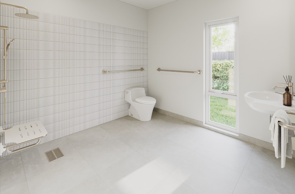 A spacious, accessible bathroom with light-coloured tiles and flooring. The room features a wall-mounted shower with a foldable seat and grab bars, a toilet with grab bars on either side, and a sink with ample space beneath it. A tall window allows natural light to enter, offering a view of greenery outside.