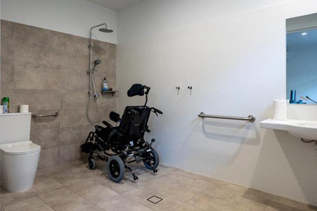 A spacious, accessible bathroom with a toilet, a wall-mounted shower, and grab bars installed for support. A specialised wheelchair is positioned near the shower area. The bathroom features a sink with ample space beneath and neutral-toned tiles on the floor and walls, creating a clean and functional space.