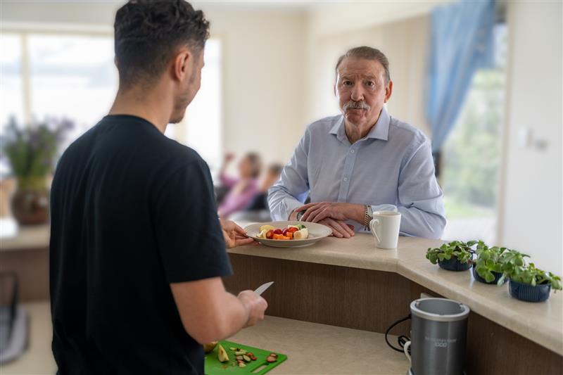 An older man in a light blue shirt stands at a kitchen counter with a plate of fruit and a cup in front of him. He faces a younger man, seen from behind, who is preparing food with a cutting board. The background shows a bright, well-lit living area with blurred details of people sitting and a window with blue curtains.
