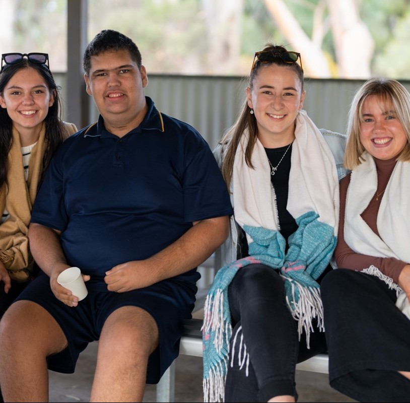 A group of four people sitting on a bench. They are smiling, and two women on the left and right are wearing light jackets, while the man in the middle is in a navy blue polo shirt. Everyone appears happy and relaxed.