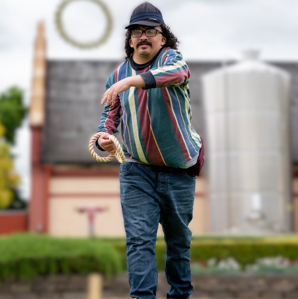 A man wearing glasses, a cap and a multi-coloured striped jumper is seen in mid-action, tossing a ring in an outdoor grassy field. In the background, there is a farm building and large metallic tanks, indicating a winery setting.