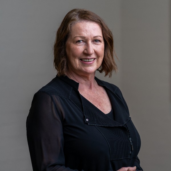 A woman with shoulder-length brown hair smiles at the camera. She is wearing a black blouse with sheer sleeves and stands against a neutral background. Her expression is friendly and confident.
