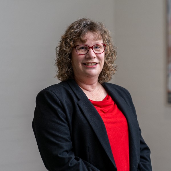 A woman with curly brown hair and red-framed glasses smiles at the camera. She is wearing a red top and a black blazer. The background is neutral, and her expression is friendly and professional.
