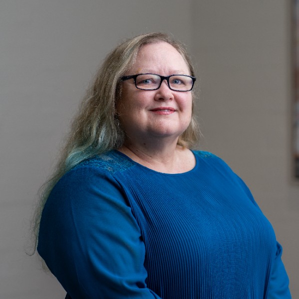 A woman with long, light brown hair that has subtle blue streaks smiles at the camera. She is wearing glasses and a bright blue blouse. The background is neutral with a blurred painting visible in the distance.