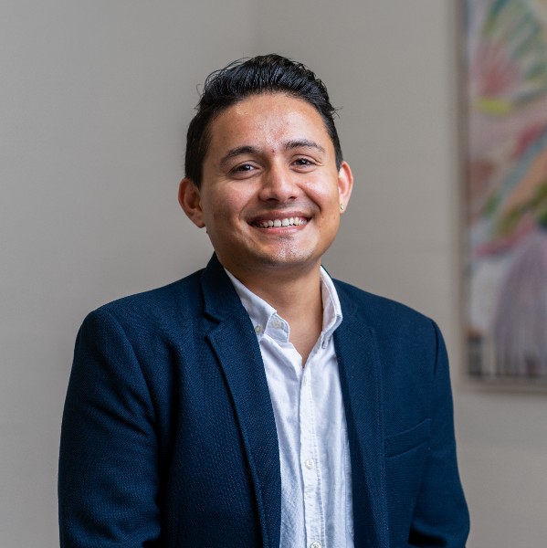 A young man with short black hair smiles warmly at the camera. He is wearing a white collared shirt and a dark blue blazer. The background is neutral with a blurred painting adding a splash of colour.