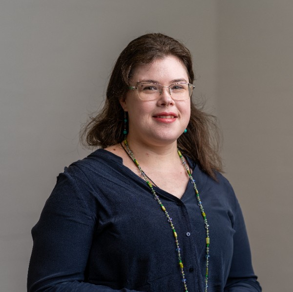 A person with shoulder-length brown hair and glasses smiles at the camera. They are wearing a dark blue top and a long beaded necklace with green and yellow accents. The background is neutral, and their expression is calm and friendly. 