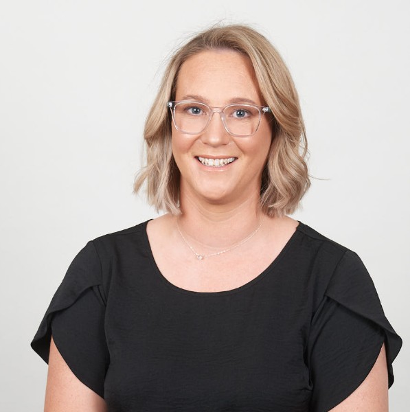 A woman with shoulder-length blonde hair and clear-framed glasses smiles at the camera. She is wearing a black short-sleeve top and a delicate necklace. The background is plain and white, giving a clean and professional appearance.
