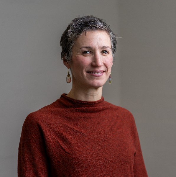 A woman with short salt-and-pepper hair smiles at the camera. She is wearing a burnt orange long-sleeve top and dangling earrings. The background is neutral, and her expression is warm and approachable.