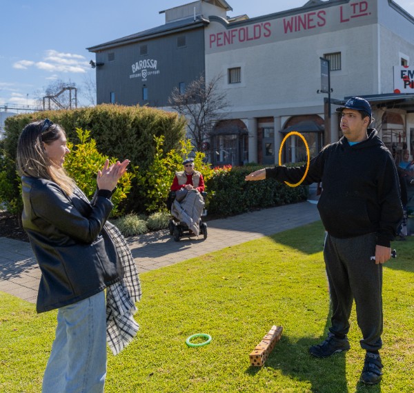 Two people engage in a game outdoors on a sunny day. One person claps encouragingly as the other balances a ring on their arm. In the background, a person in a wheelchair observes the activity.