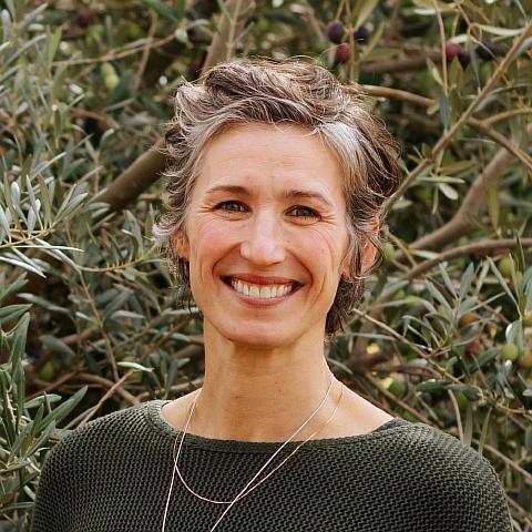 A woman with short salt-and-pepper hair smiles at the camera. She is wearing a burnt orange long-sleeve top and dangling earrings. The background is neutral, and her expression is warm and approachable.