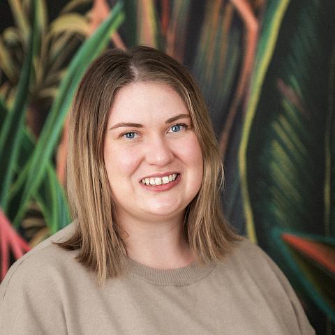 A woman with shoulder-length wavy brown hair smiles at the camera. She is wearing a dark green blouse and a delicate necklace. The background is neutral, and her expression is warm and friendly
