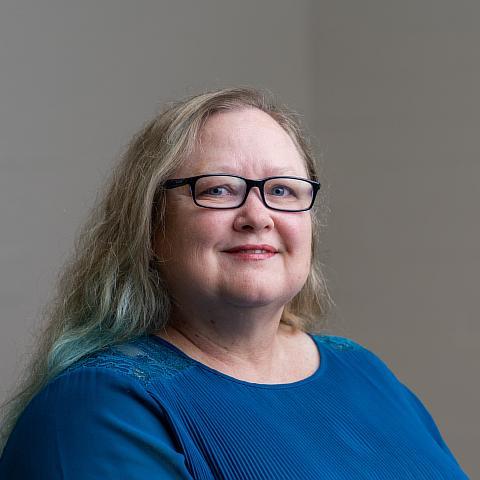 A woman with long, light brown hair that has subtle blue streaks smiles at the camera. She is wearing glasses and a bright blue blouse. The background is neutral with a blurred painting visible in the distance.