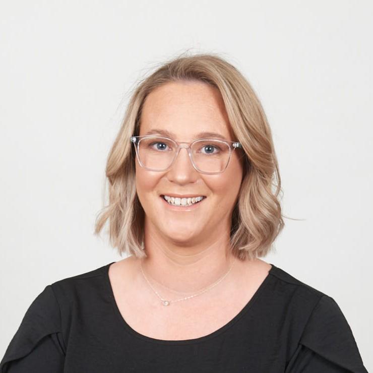 A woman with shoulder-length blonde hair and clear-framed glasses smiles at the camera. She is wearing a black short-sleeve top and a delicate necklace. The background is plain and white, giving a clean and professional appearance.