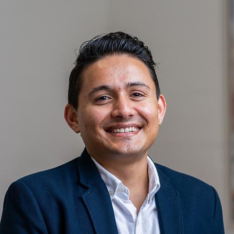 A young man with short black hair smiles warmly at the camera. He is wearing a white collared shirt and a dark blue blazer. The background is neutral with a blurred painting adding a splash of colour.
