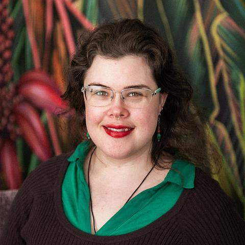 A person with shoulder-length brown hair and glasses smiles at the camera. They are wearing a dark blue top and a long beaded necklace with green and yellow accents. The background is neutral, and their expression is calm and friendly. 