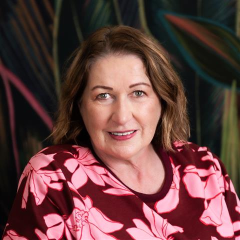 A woman with shoulder-length brown hair smiles at the camera. She is wearing a black blouse with sheer sleeves and stands against a neutral background. Her expression is friendly and confident.