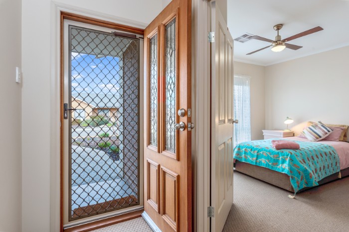A well-lit entryway with a wooden front door and a screen door, leading outside. To the right of the entryway is a bedroom with a neatly made bed, featuring a teal and pink patterned blanket. A ceiling fan and bedside lamp are visible in the bedroom.