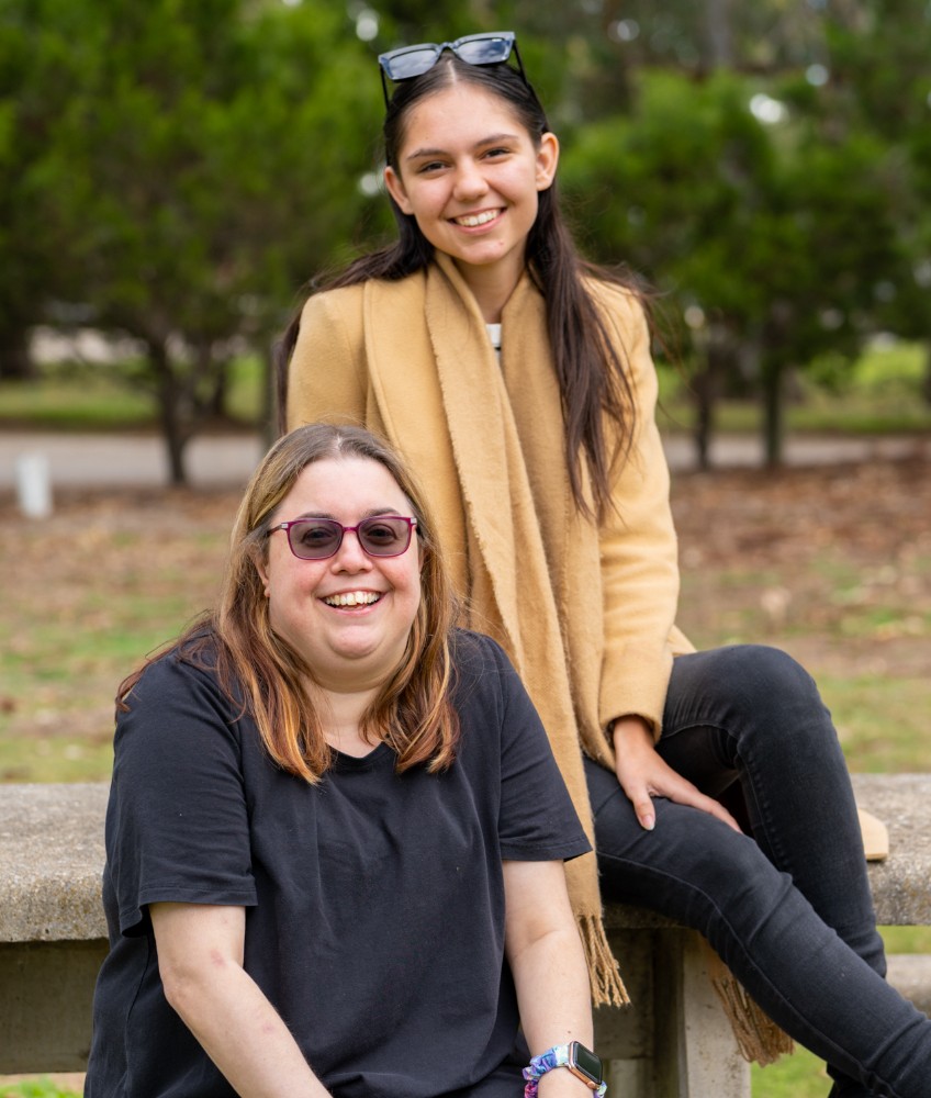 Two women smiling outdoors, sitting on a stone bench in a park. The woman in front, wearing a black t-shirt and sunglasses, is seated, while the woman behind her, wearing a tan coat and sunglasses on her head, is perched on the bench. The background features trees and greenery.