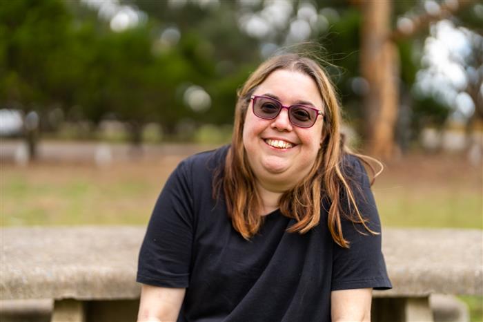 A smiling woman with long light brown hair wearing sunglasses and a black t-shirt sits outdoors on a stone bench. The background is a park-like setting with blurred greenery and trees.