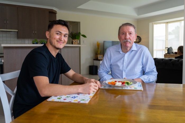 Two men sit at a dining table in a home kitchen. The younger man on the left, wearing a black t-shirt, smiles at the camera. The older man on the right, wearing a light blue shirt, has a neutral expression. A plate of food with vegetables is in front of the older man. The background shows a modern kitchen and living room.