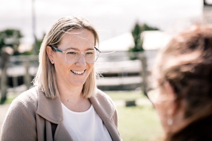 A woman with blonde hair, wearing glasses and a beige coat, smiles while speaking to another person whose back is to the camera. They are outdoors in a sunny setting with blurred greenery and fencing in the background.