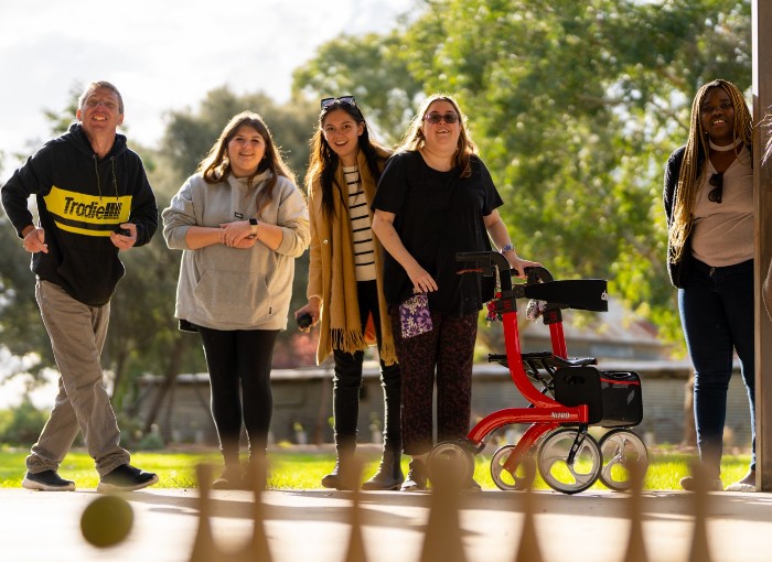 A group of five people stand outdoors, smiling and engaging in a bowling game. One person uses a walker for support, and another holds a ball, preparing to bowl. They appear to be enjoying a casual, fun activity in a park-like setting with trees and greenery in the background.