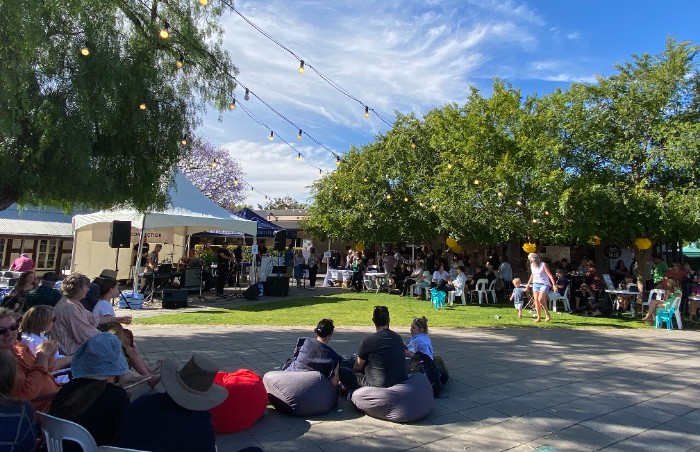A lively outdoor event with people sitting on chairs and bean bags, enjoying live music performed on a stage under a tent. String lights are hanging above, and the area is shaded by trees. A mix of people are seated and standing, with others walking around the grassy and paved areas. The sky is clear with scattered clouds, adding to the festive atmosphere
